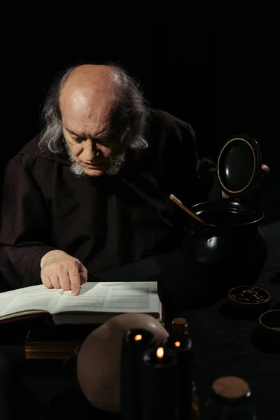 Senior alchemist reading magic cookbook near pot isolated on black — Stock Photo