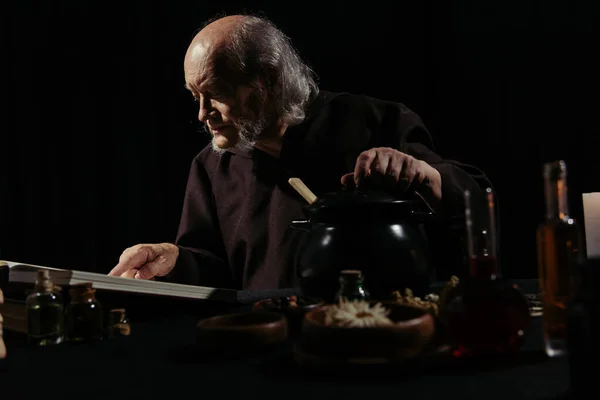 Medieval wizard reading magic cookbook near pot and ingredients isolated on black — Stock Photo