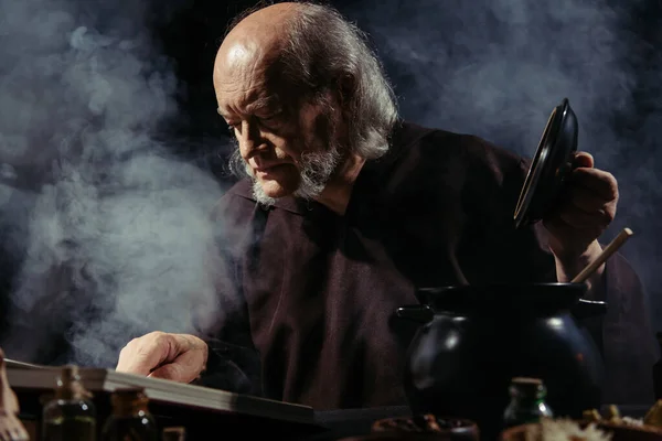 Alquimista medieval lendo livro de receitas mágico enquanto prepara poção à noite em fundo preto com fumaça — Fotografia de Stock