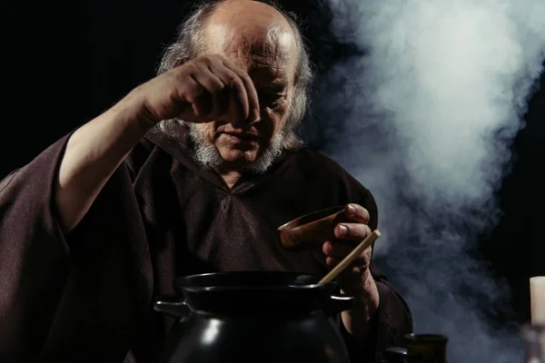 Mago anziano aggiungendo ingrediente dalla ciotola in pentola su sfondo nero con fumo — Foto stock