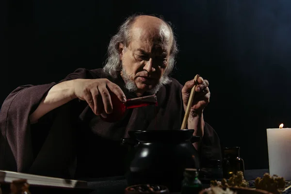 Mysterious alchemist preparing magic potion and pouring liquid into pot isolated on black — Stock Photo