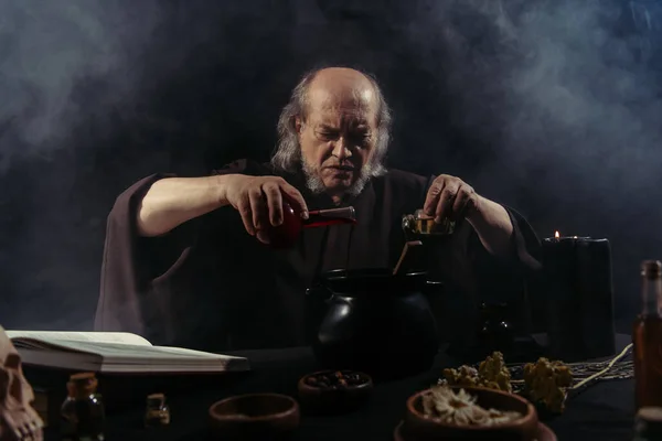 Senior magician pouring liquid into pot while preparing potion on black background with smoke — Stock Photo