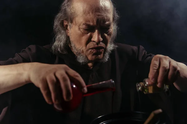 Alquimista barbudo mezclando ingredientes líquidos y preparando poción mágica aislada en negro - foto de stock