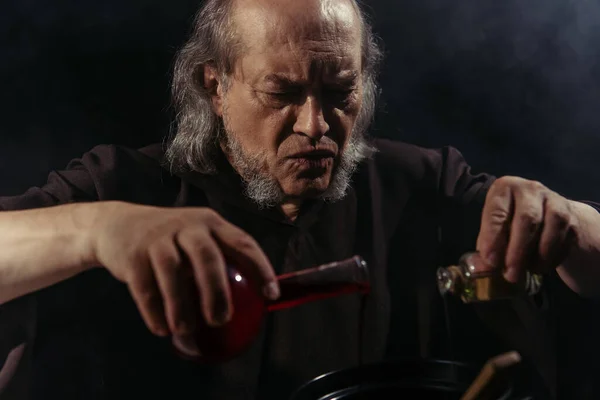 Bearded alchemist mixing liquid ingredients and preparing magic potion isolated on black — Stock Photo