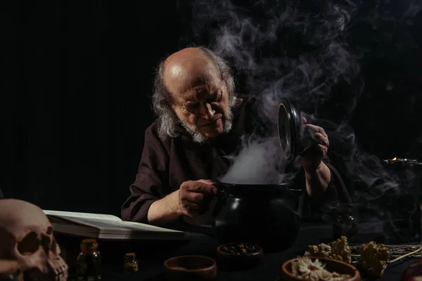 Medieval alchemist near boiling pot and magic cookbook on black background — Stock Photo