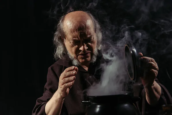Alchimista anziano accigliato vicino pentola fumante isolato su nero — Foto stock