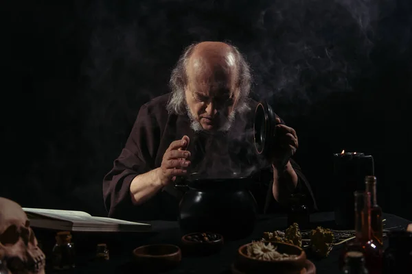 Alchemist in dark robe looking into steaming pot near herbal ingredients on black background — Stock Photo