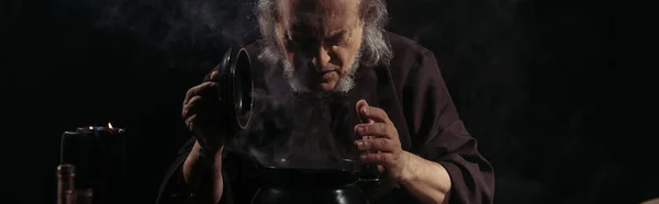 Senior alchemist looking in boiling pot with potion isolated on black, banner — Stock Photo