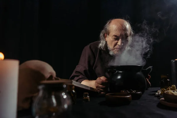 Alquimista senior cerca de hervir y olla al vapor preparando poción en la noche aislado en negro - foto de stock
