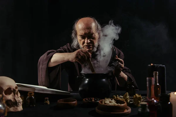 Senior wizard mixing boiling potion in steaming pot at night isolated on black — Stock Photo