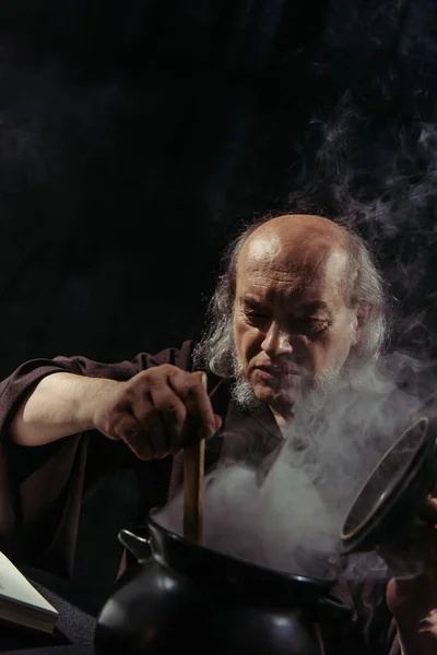 Средневековый целитель готовит волшебное зелье в кипящем котле, изолированном на черном — стоковое фото