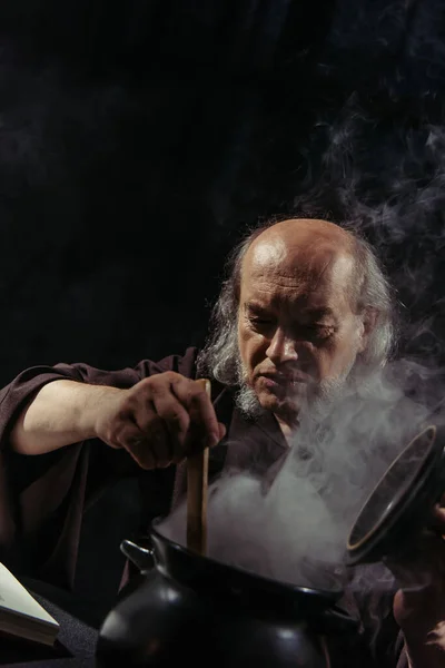 Medieval healer preparing magic potion in boiling pot isolated on black — Stock Photo