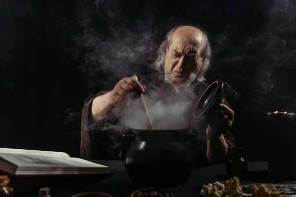 Alquimista senior frunciendo el ceño cerca de olla humeante mientras cocina por la noche sobre fondo negro - foto de stock
