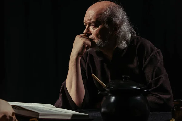 Serious alchemist thinking near magic cookbook and pot isolated on black — Stock Photo