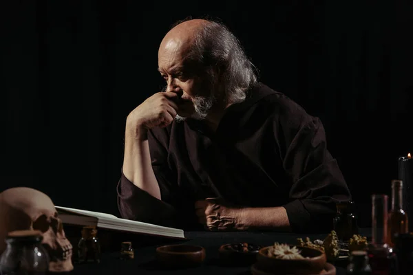 Alquimista reflexivo lectura libro de cocina mágica cerca de hierbas secas aisladas en negro - foto de stock