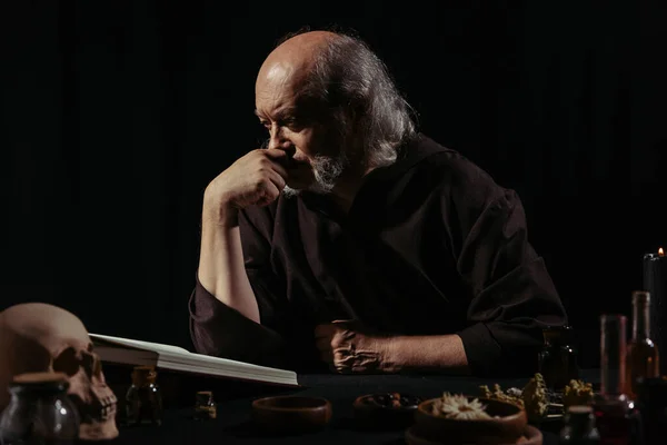 Thoughtful alchemist reading magic cookbook near dried herbs isolated on black — Stock Photo