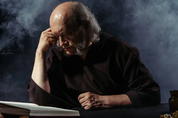 Alquimista senior leyendo libro de cocina mágica en la noche sobre fondo negro con humo - foto de stock