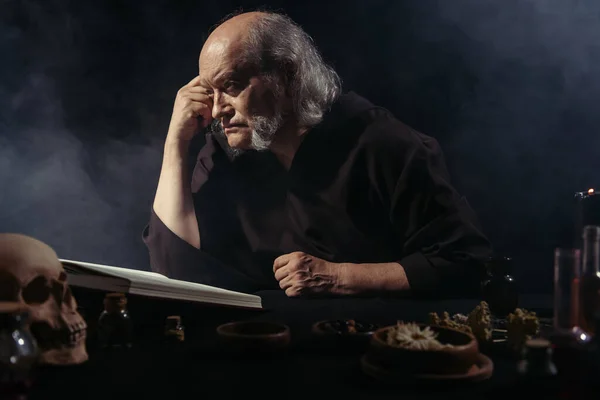 Medieval alchemist thinking near magic cookbook and dried herbs on black background with smoke — Stock Photo