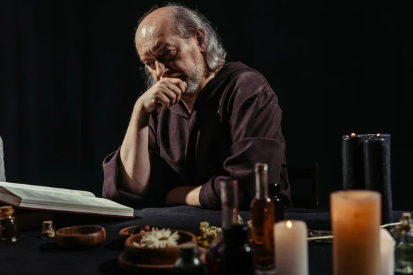Alchimiste réfléchi lecture livre de cuisine magique près des ingrédients et des bougies isolées sur noir — Photo de stock