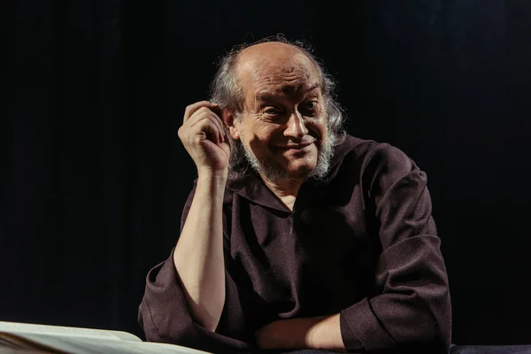 Smiling monk looking at camera while sitting near bible isolated on black — Stock Photo