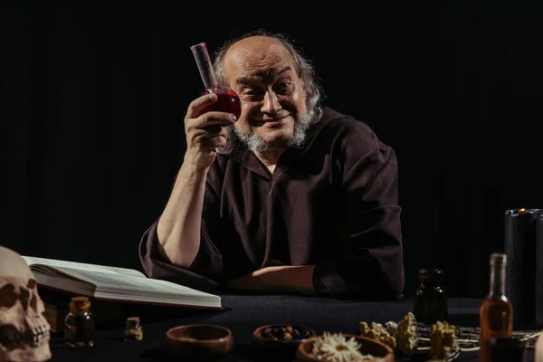 Alchimiste joyeux montrant fiole avec potion près de livre de cuisine magique isolé sur noir — Photo de stock