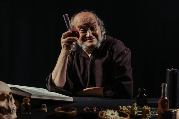 Cheerful alchemist showing flask with potion near magic cookbook isolated on black — Stock Photo