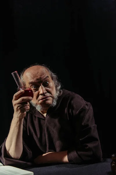 Excited alchemist holding flask with magic potion and looking at camera isolated on black — Stock Photo