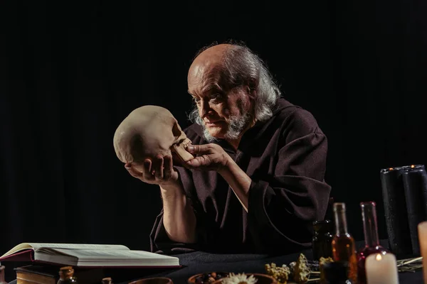 Mysterious alchemist looking at human skull near magic cookbook and dried herbs isolated on black — Stock Photo