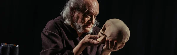Medieval philosopher with grey hair looking at human skull isolated on black, banner — Stock Photo