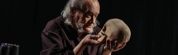 Filósofo medieval con el pelo gris mirando cráneo humano aislado en negro, bandera - foto de stock