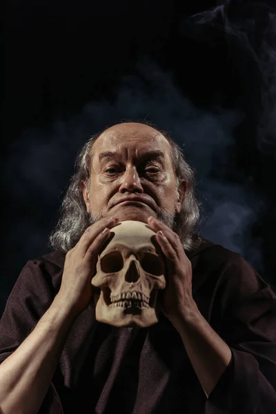 Thoughtful religious philosopher looking at camera near human skull on black background — Stock Photo