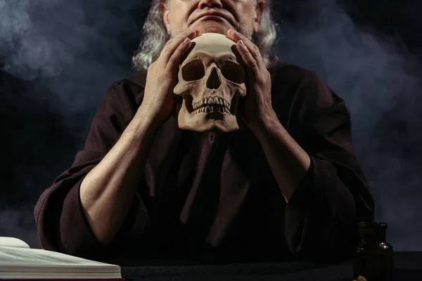 Cropped view of medieval alchemist with skull sitting on black background with smoke — Stock Photo