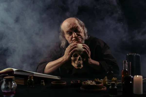 Discouraged alchemist holding skull near ingredients and magic cookbook on black background with smoke — Stock Photo