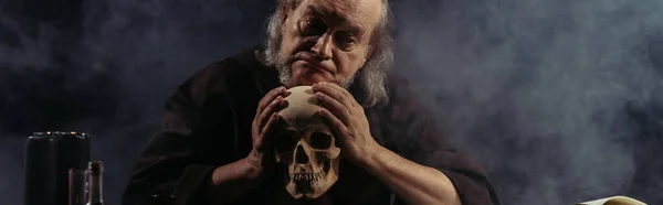 Thoughtful senior wizard holding human skull on black background with smoke, banner — Stock Photo