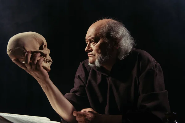 Senior philosopher in dark robe looking at human skull isolated on black — Stock Photo