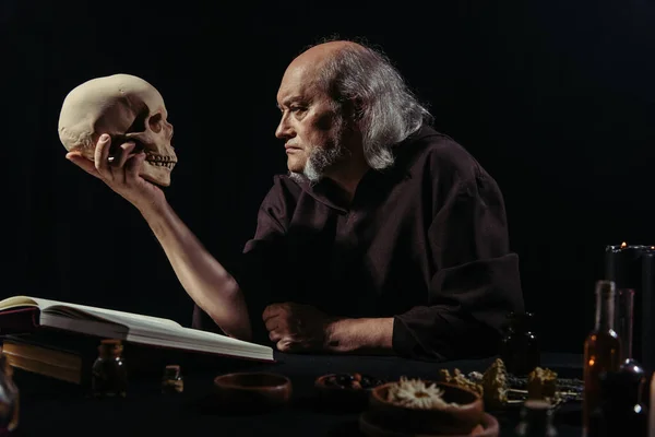 Senior alchemist looking at skull near magic cookbook and herbal ingredients isolated on black — Stock Photo