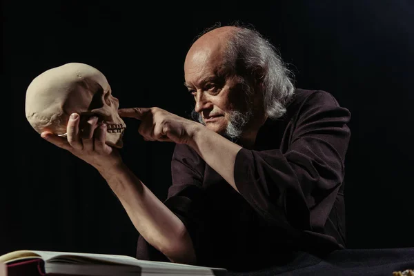 Senior medieval philosopher pointing at human skull isolated on black — Stock Photo