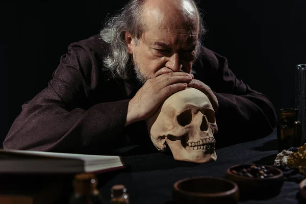 Старший алхімік мислення біля черепа і миски з сушеними травами ізольовані на чорному — стокове фото