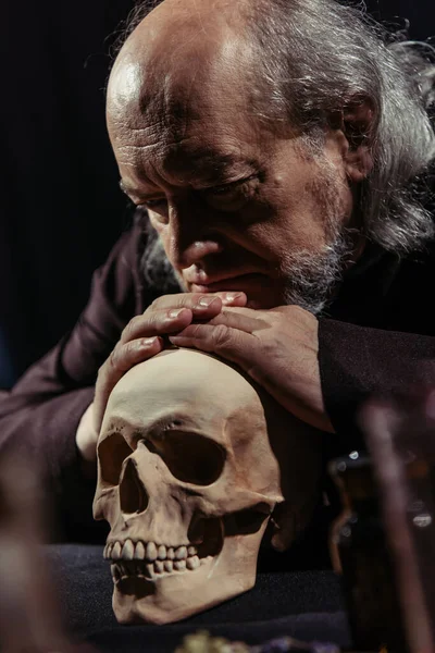 Senior medieval philosopher leaning on skull while thinking isolated on black — Stock Photo