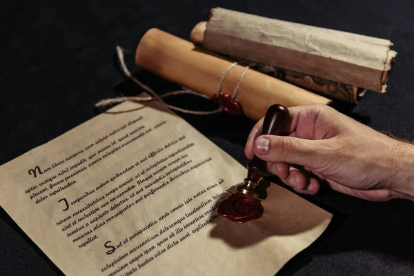 Rolled parchments near cropped monk holding wax seal near ancient manuscript on black surface — Stock Photo