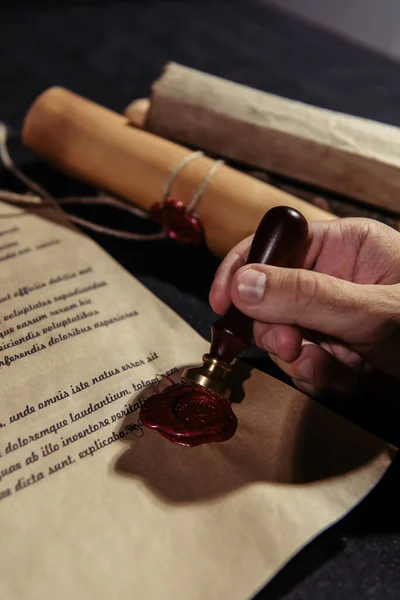 Vista recortada del sacerdote con sello de cera cerca de pergaminos laminados y manuscrito antiguo en la superficie negra - foto de stock