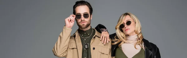 Stylish man adjusting sunglasses near blonde woman in leather jacket isolated on grey, banner — Stockfoto