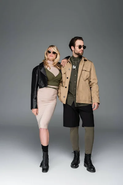 Full length of stylish couple in autumnal outfits and sunglasses posing together on grey - foto de stock