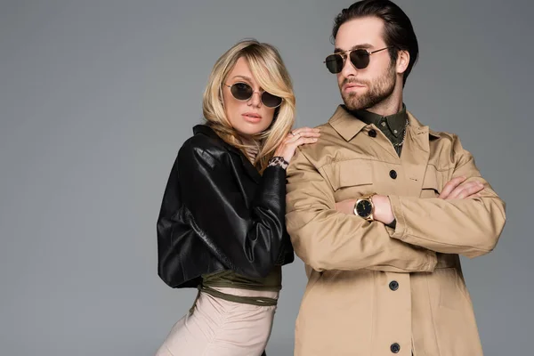 Stylish man in sunglasses posing with crossed arms near blonde woman in leather jacket and dress isolated on grey - foto de stock