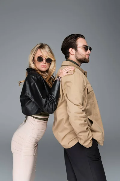 Stylish blonde woman in sunglasses leaning on shoulders on bearded man isolated on grey - foto de stock