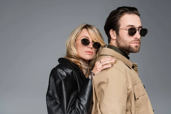 Stylish blonde woman in sunglasses and leather jacket leaning on shoulders on bearded man isolated on grey — Stockfoto