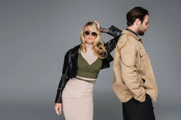 Stylish blonde woman in sunglasses and leather jacket leaning on back of bearded man isolated on grey — Stockfoto