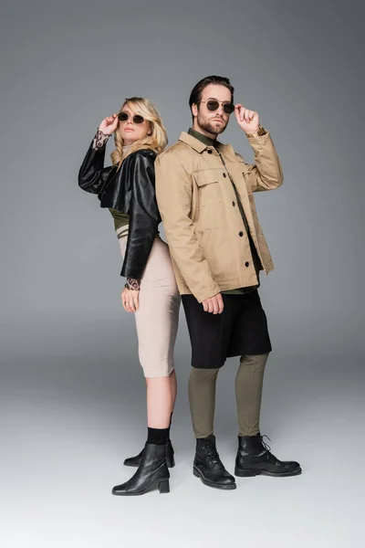 Full length of stylish couple in autumnal outfits adjusting sunglasses and posing together on grey — Foto stock