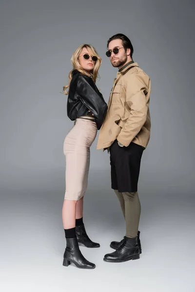 Full length of stylish couple in trendy outfits and sunglasses posing together on grey — Stock Photo