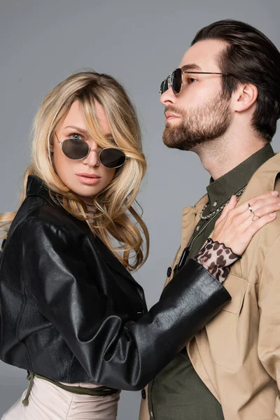Stylish blonde in black jacket woman hugging bearded man in sunglasses isolated on grey — Photo de stock